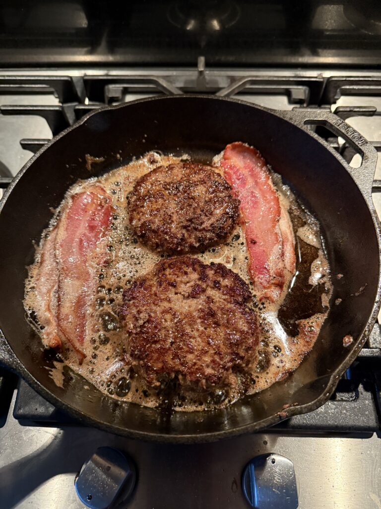 Twee sappige runderburgers en plakken bacon die samen braden in een gietijzeren pan.